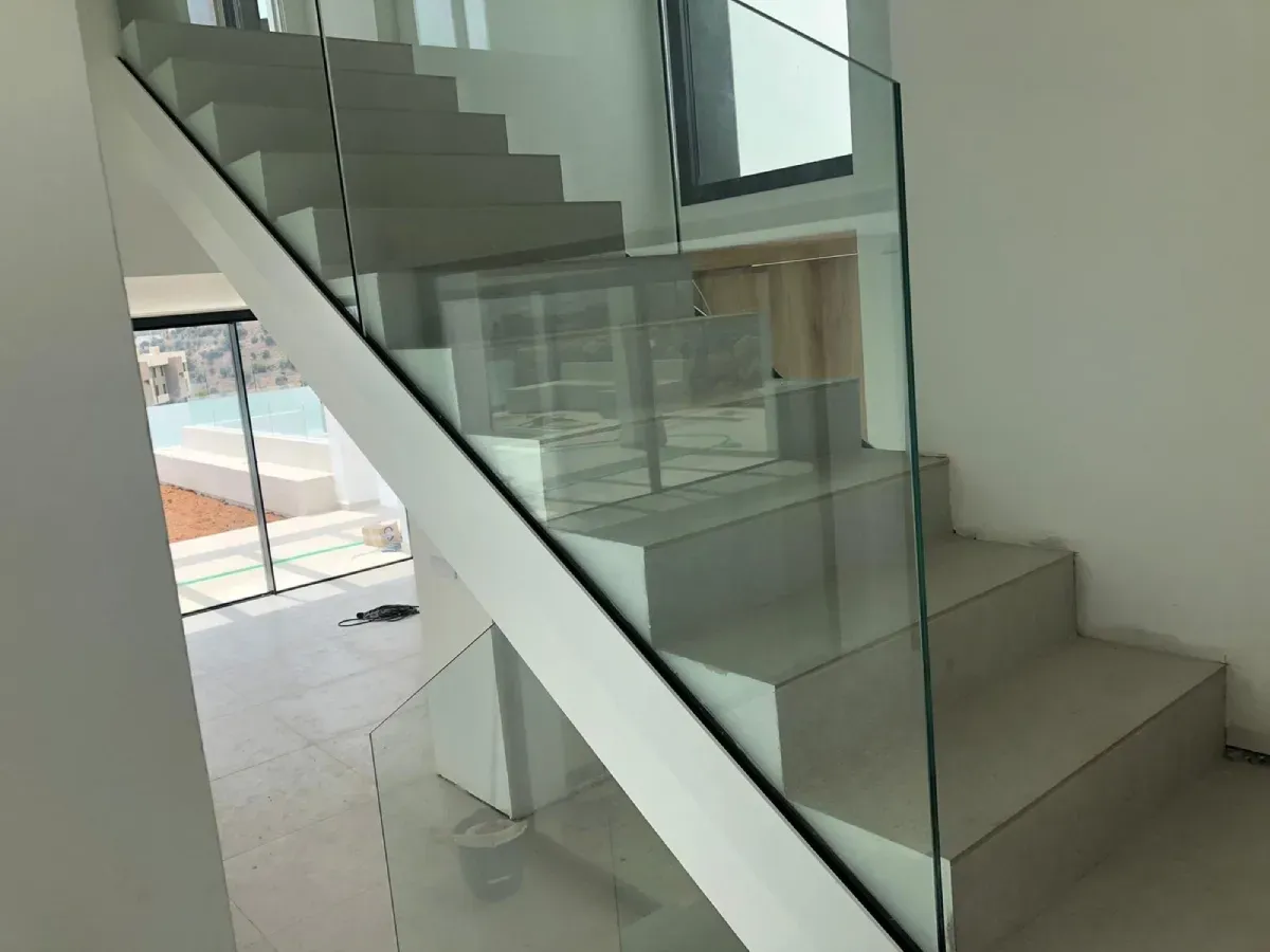 Escalera interior minimalista con escalones de color claro y barandilla de cristal sin marco, vista desde un lateral.