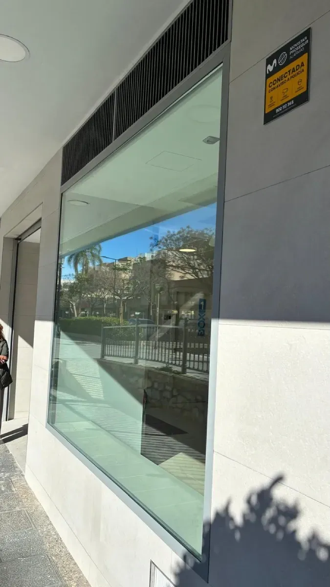 Un gran escaparate con superficie reflectante, paredes grises y blancas, y un cartel de sistema de seguridad en la esquina.