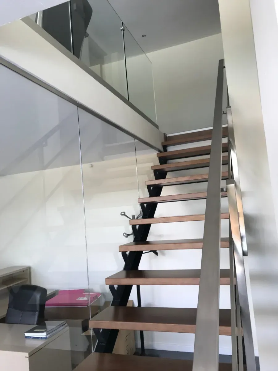 una escalera con peldaños de madera oscura y estructura de metal negro conduce a un nivel superior con barandilla de cristal.