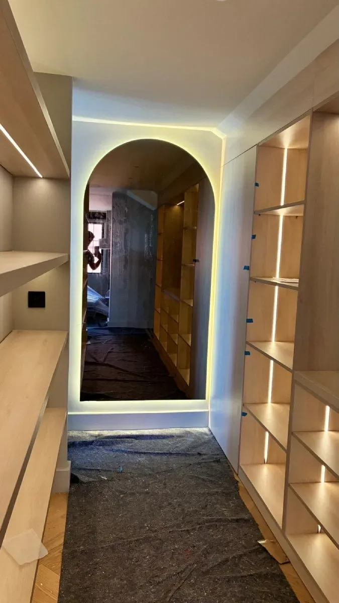 Un vestidor en construcción con estanterías de madera, iluminación LED de acento y un espejo arqueado al fondo.