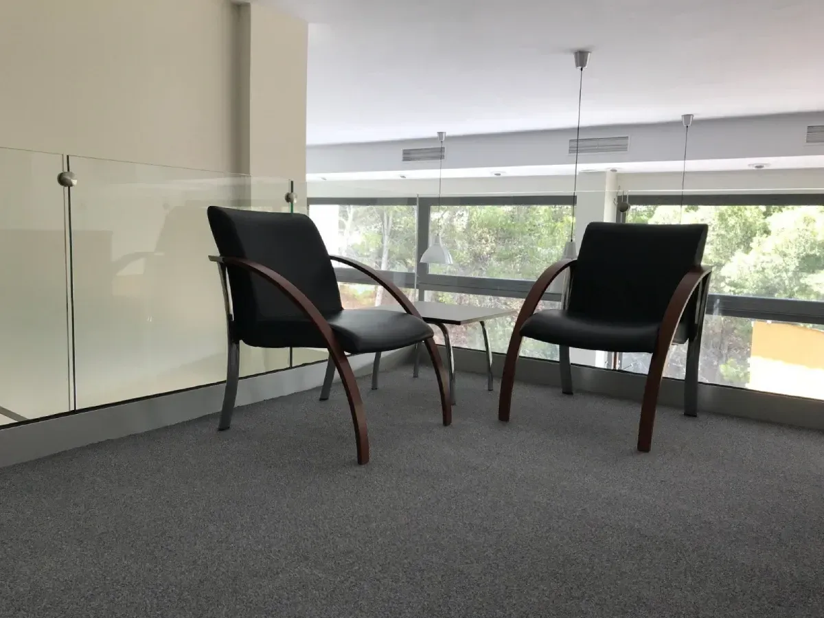 Dos sillas negras con reposabrazos de madera en un rellano alfombrado de color gris, junto a una barandilla de vidrio.