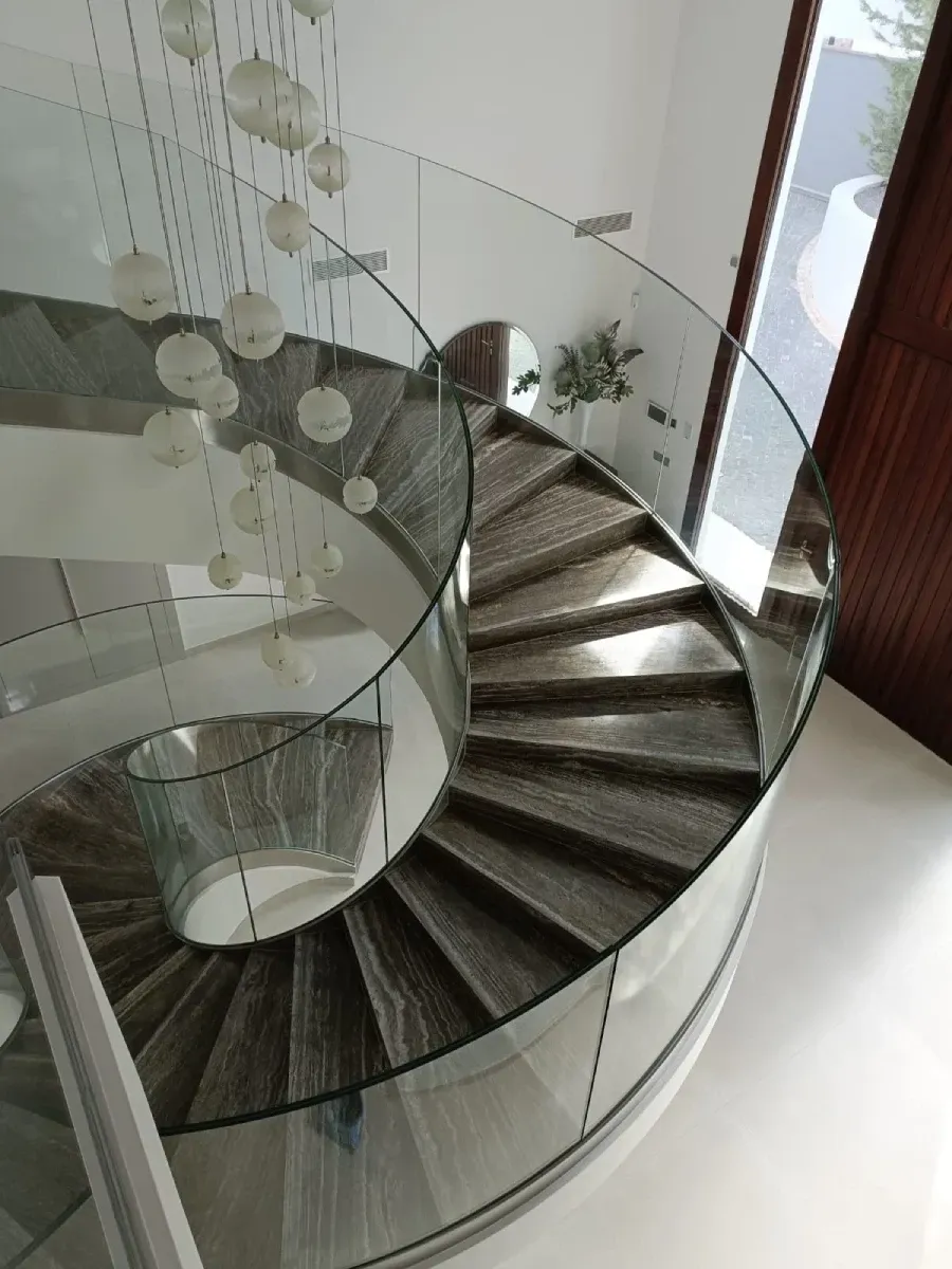 Vista desde un ángulo elevado de una escalera de caracol con escalones de madera y una barandilla de cristal transparente.
