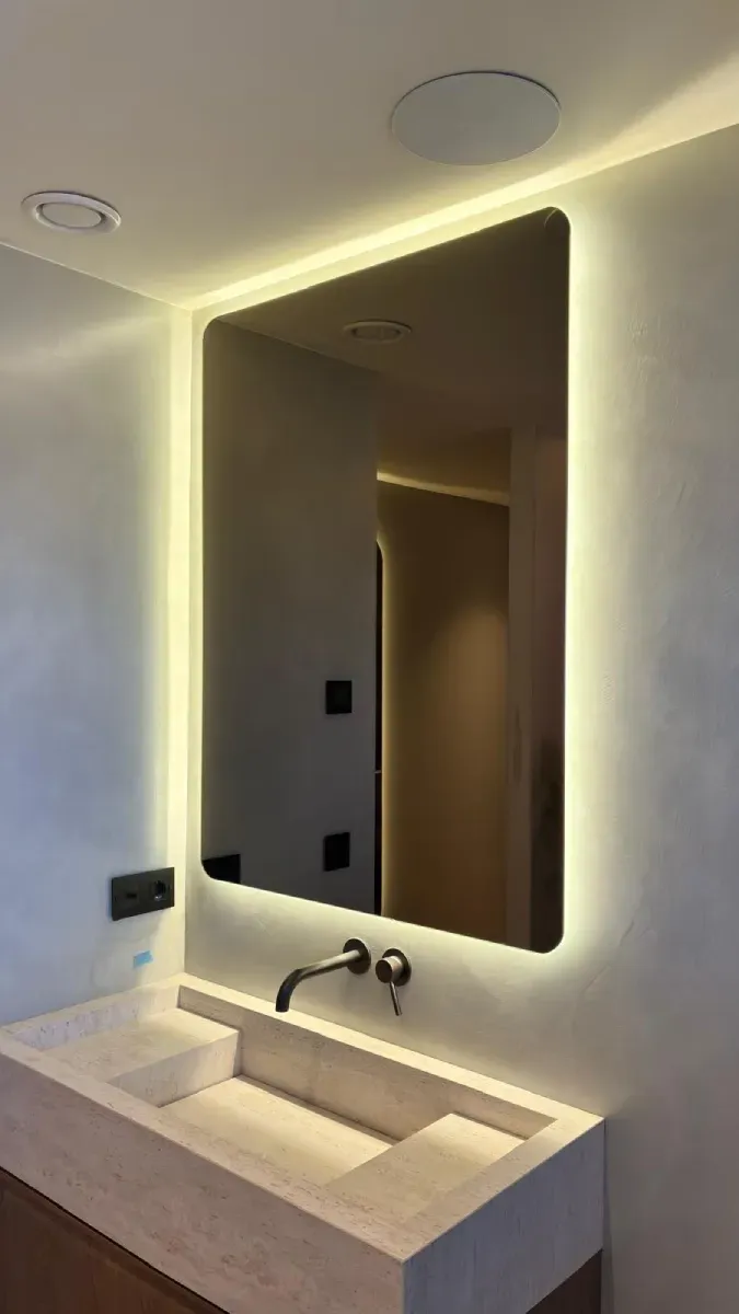Tocador de baño moderno con espejo rectangular retroiluminado sobre lavabo de piedra y grifo mural.