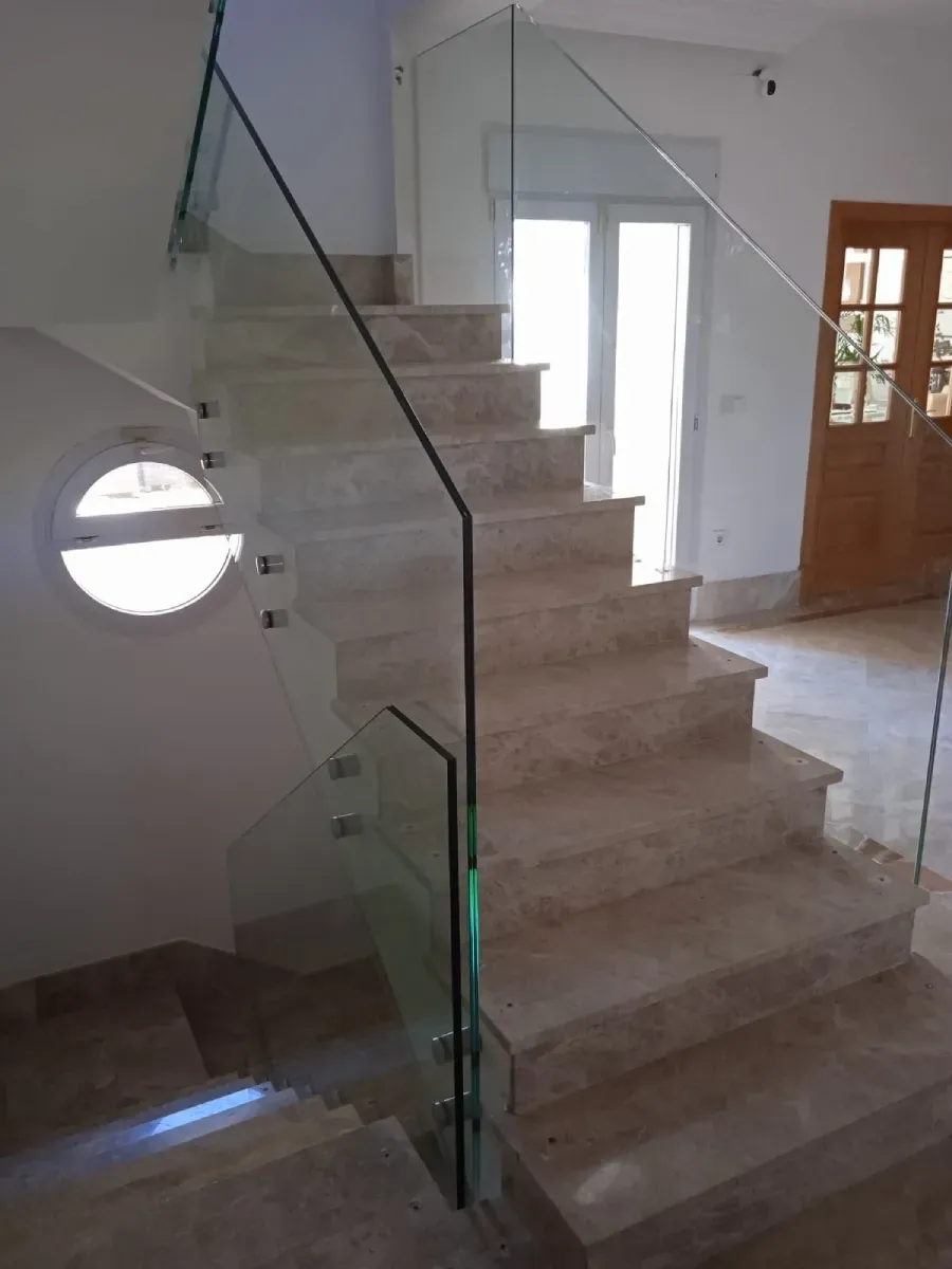 Una escalera de mármol, con una barandilla de vidrio con soportes metálicos y una ventana circular.