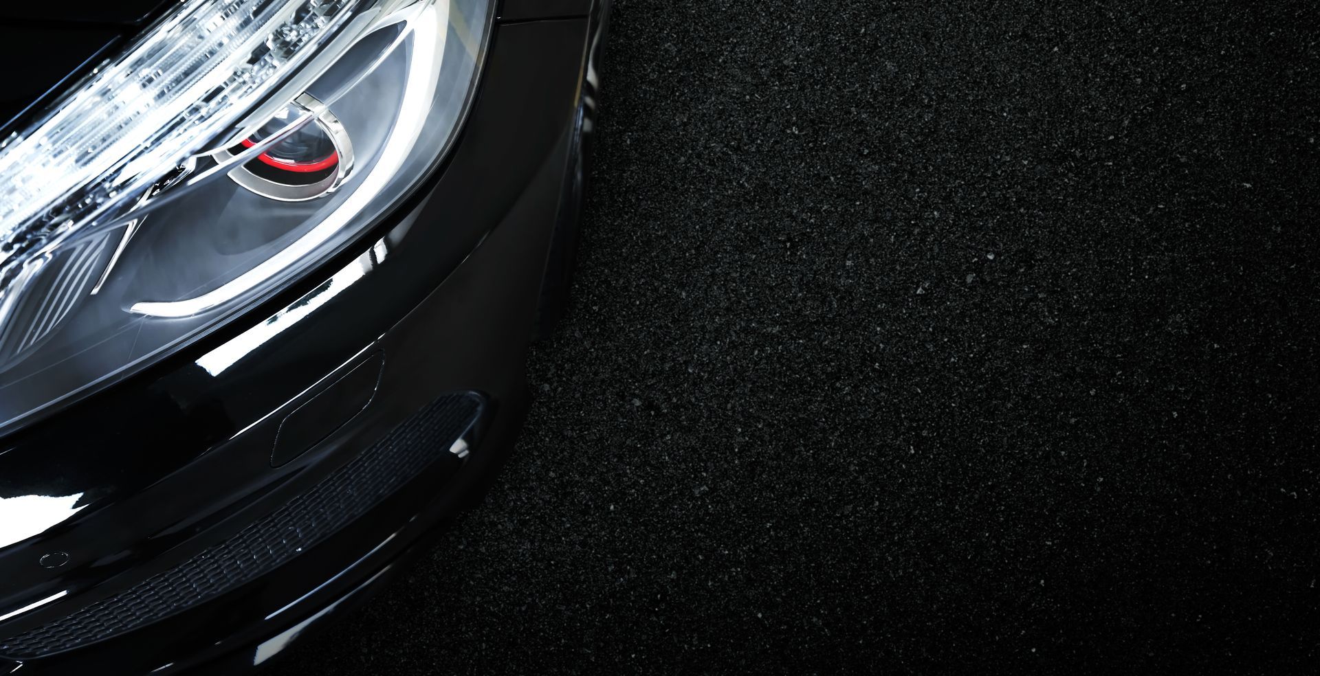 Gros plan sur le phare et le pare-chocs avant d'une voiture de sport noire sur une surface sombre et texturée.