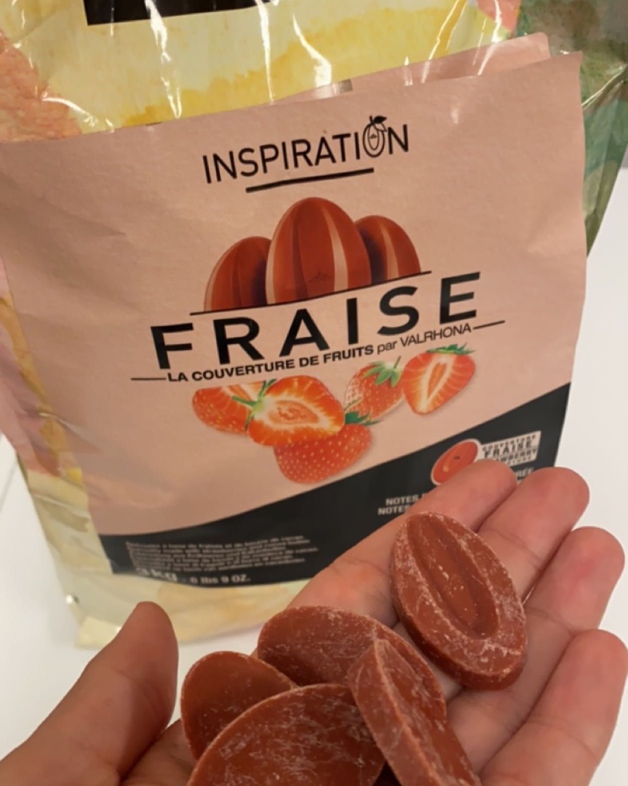 Main tenant des disques de chocolat à la fraise, avec un sachet de couverture de fruits Valrhona FRAISE en arrière-plan.