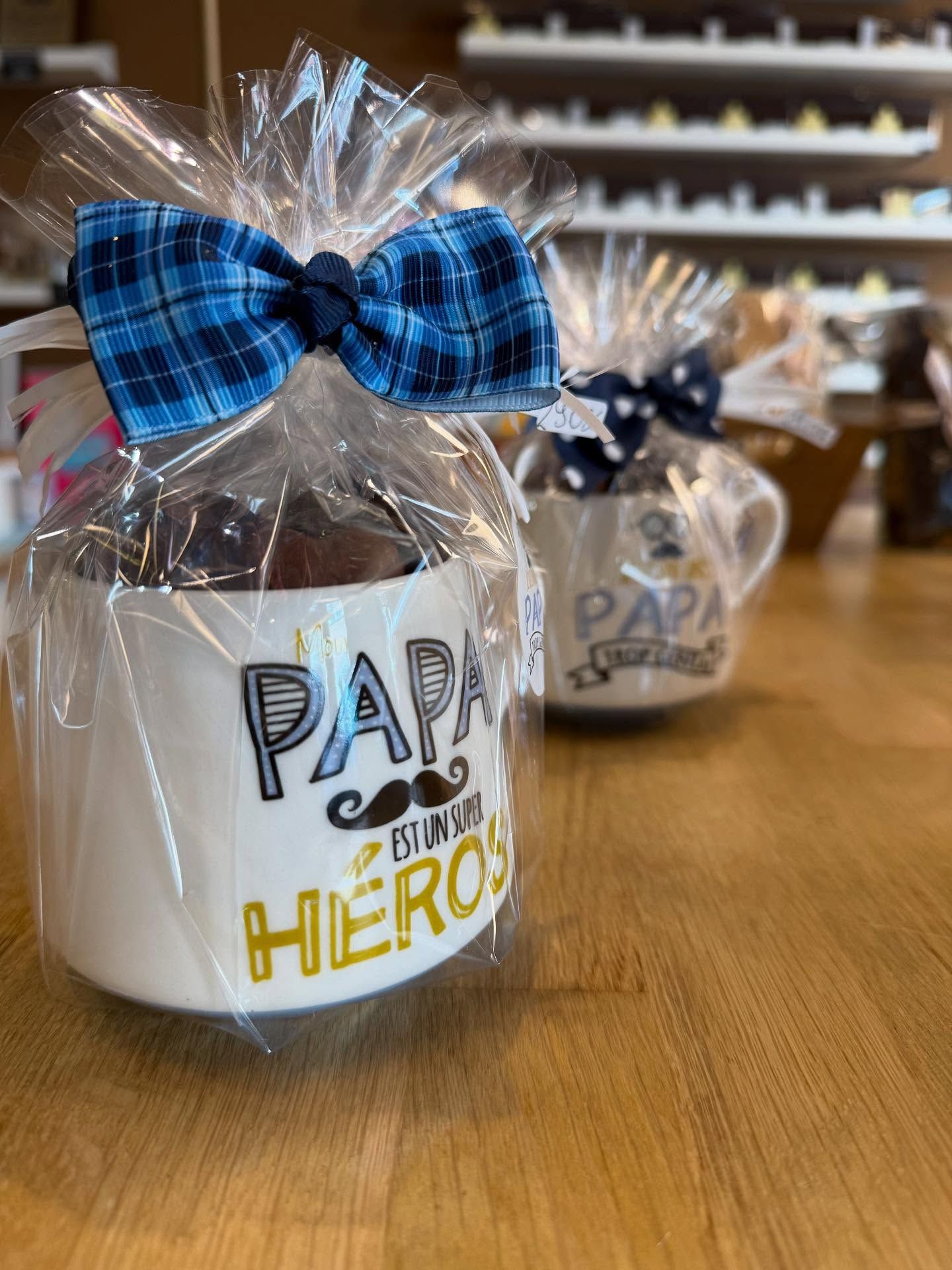 Deux mugs emballés sur une surface en bois. L'un porte l'inscription PAPA est un superhéros, avec une moustache dessinée, l'autre PAPA trop génial.