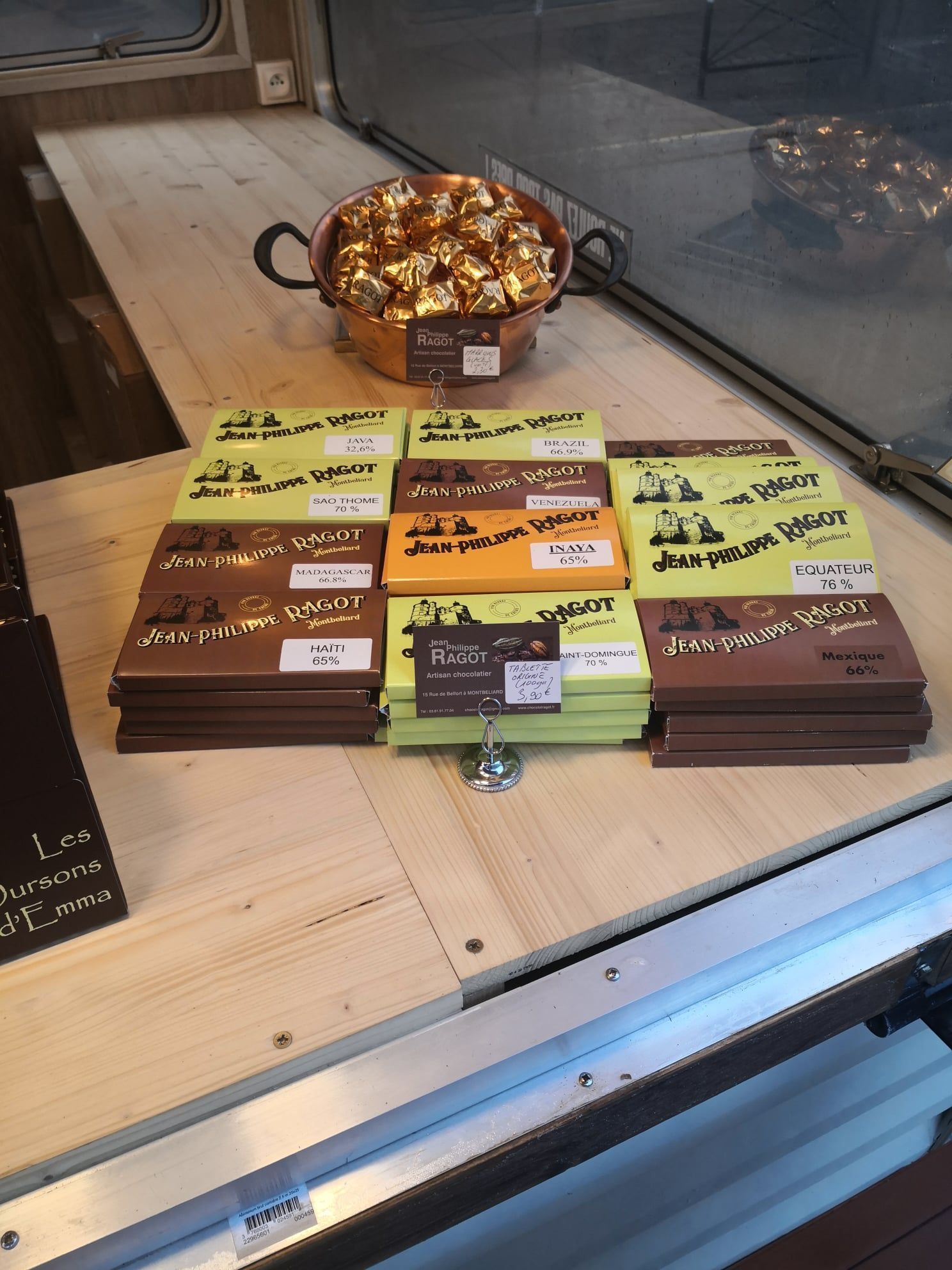 Exposition de barres de chocolat et de bonbons emballés dans du papier doré dans un magasin, sur une surface en bois derrière une vitre.