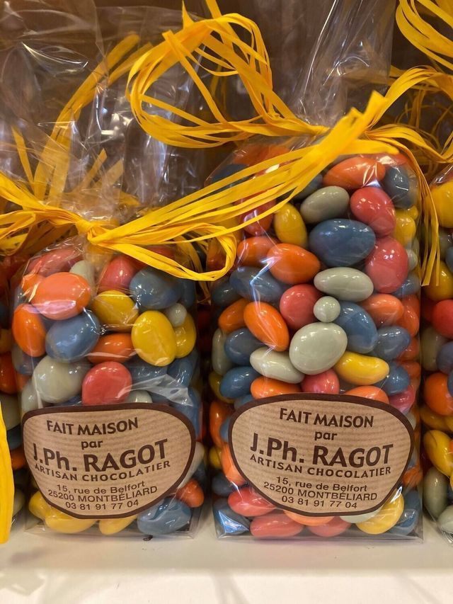 Des sacs de bonbons colorés enrobés, noués avec un ruban jaune, étiquetés J.Ph. Ragot, Artisan Chocolatier.
