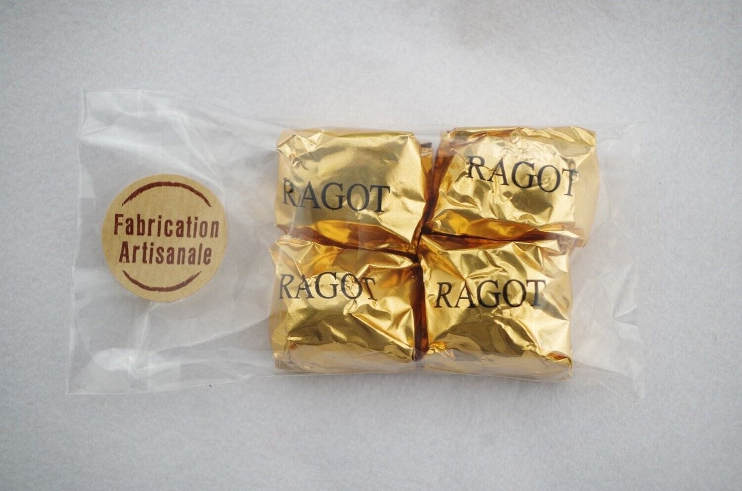 Quatre carrés de chocolat enveloppés doré dans un sachet en plastique transparent avec une étiquette Fabrication Artisanale.