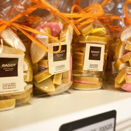 Des sachets de bonbons colorés, attachés avec des rubans orange, sur une étagère avec des étiquettes RAGOT.