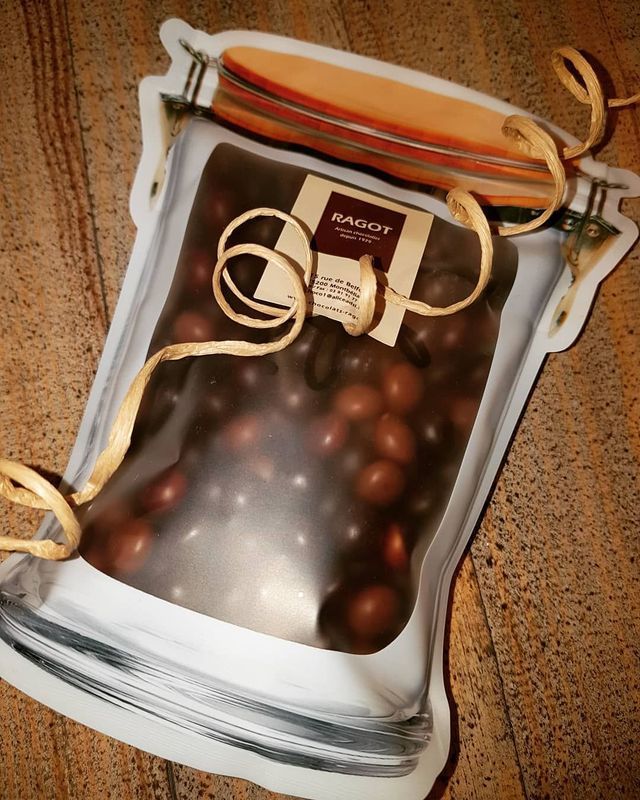 Sachet en forme de pot contenant des bonbons enrobés de chocolat. Bonbons marron à l'intérieur, ruban et étiquette beiges.