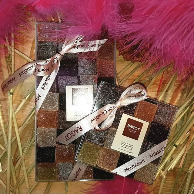 Deux boîtes de pâtes de fruits colorées Ragot Artisan Chocolat, nouées avec des rubans, posées sur des plumes roses et de la paille.