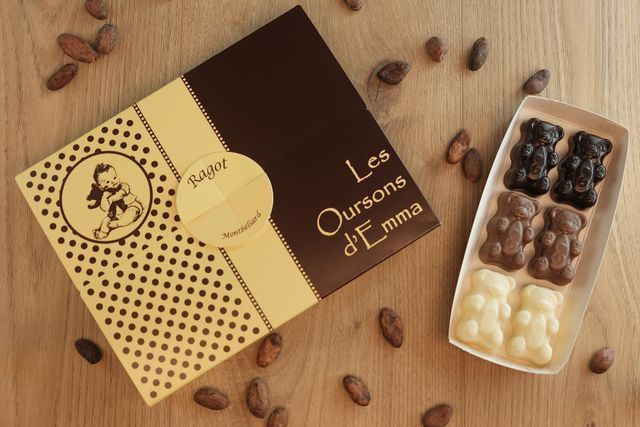 Boîte de chocolats Les Oursons d'Emma, ouverte pour révéler des bonbons en forme d'oursons. Des fèves de cacao l'entourent sur une table en bois.