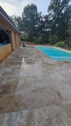 Terrasse en béton jouxtant une piscine turquoise, avec des arbres et un bâtiment en arrière-plan.