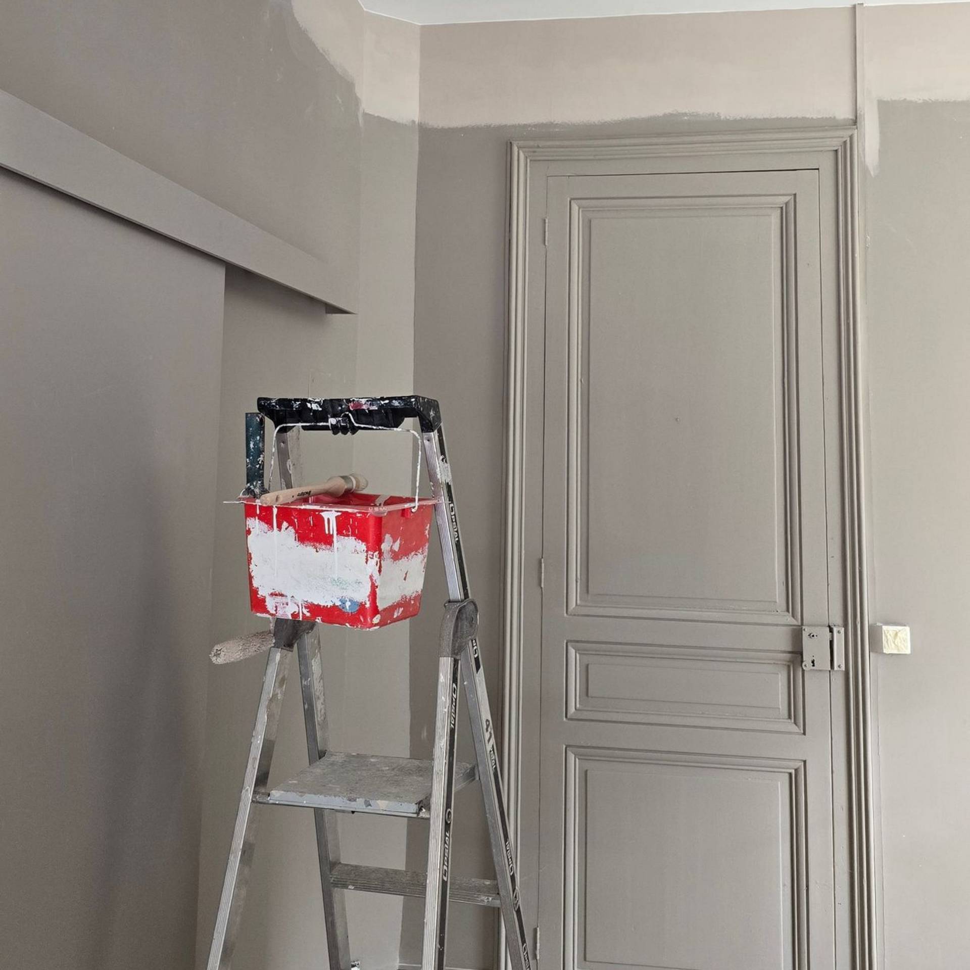 Un pot de peinture sur un escabeau dans une pièce grise aux portes à panneaux fermées.