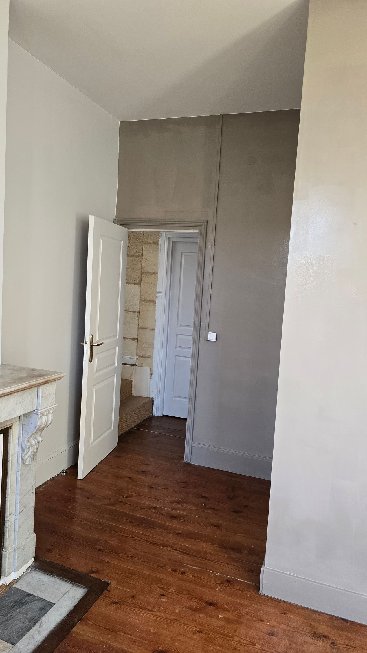 Couloir étroit aux murs blancs, porte ouverte, mur d'accent gris et parquet foncé.