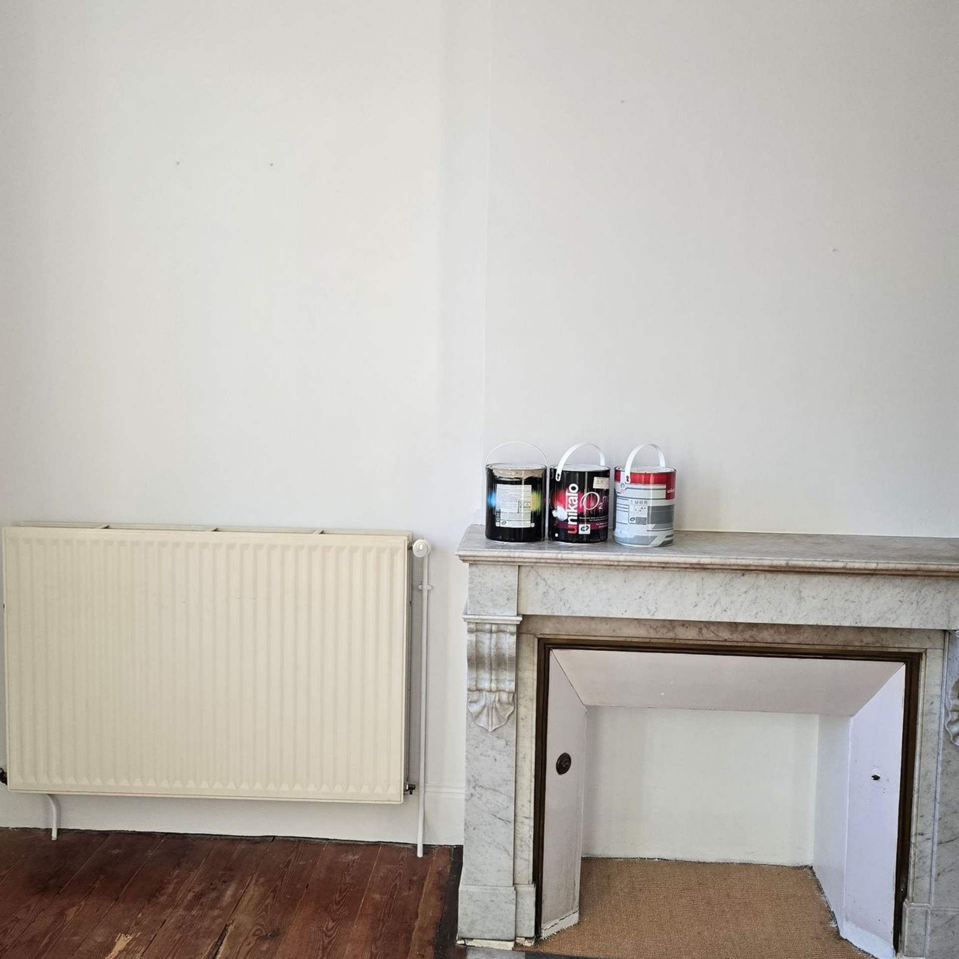 Pièce vide aux murs blancs, avec un radiateur couleur crème et des pots de peinture sur une cheminée.