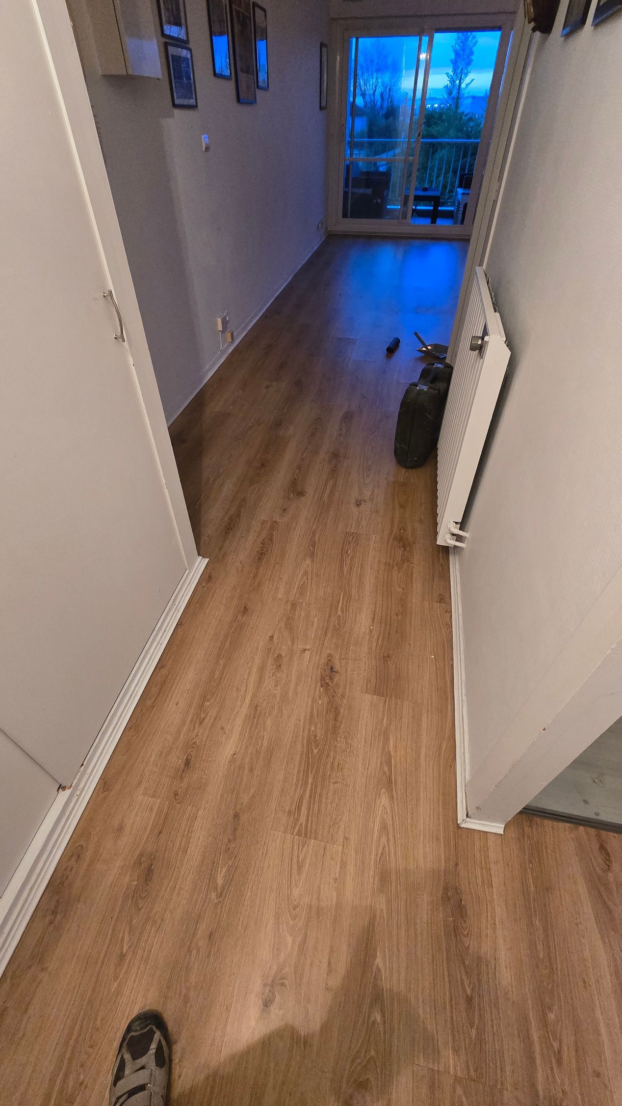 Couloir étroit avec parquet, portes ouvertes, radiateur et une pièce éclairée en bleu au fond.