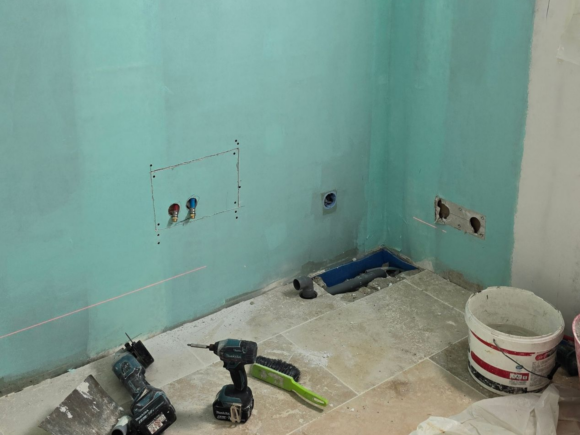 Pièce partiellement rénovée avec des murs verts, des outils au sol et un seau près des prises électriques.