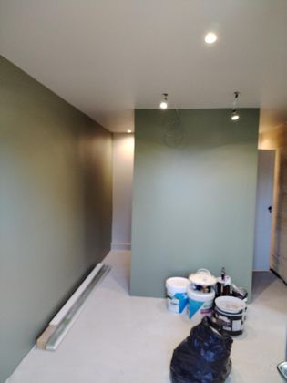 Pièce en rénovation avec des murs verts et blancs, des seaux de peinture, des outils et une bâche noire au sol.