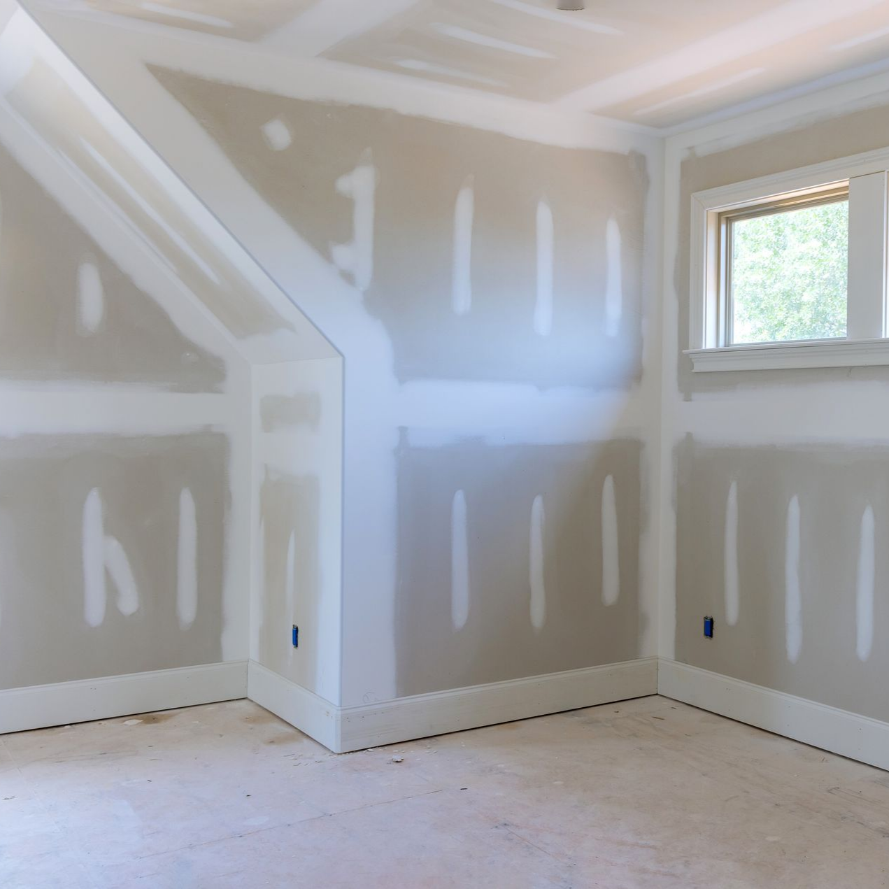 Pièce vide et inachevée avec des cloisons sèches grises, des moulures blanches et une petite fenêtre.