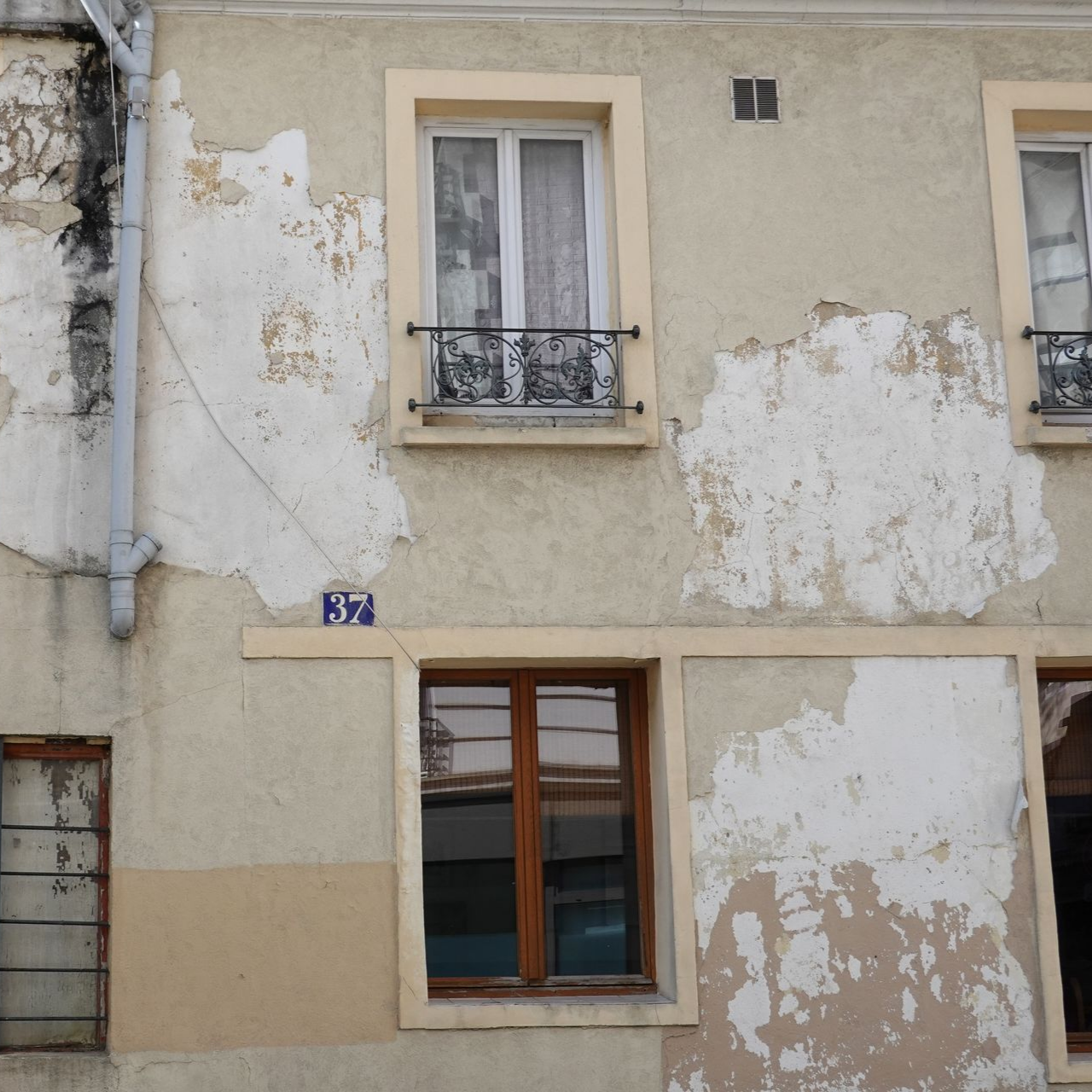 Façade d'appartement beige défraîchie, peinture écaillée, deux fenêtres et un petit balcon.