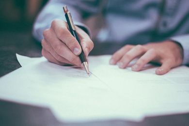 Primer plano de unas manos escribiendo con un bolígrafo sobre papel blanco en un escritorio.