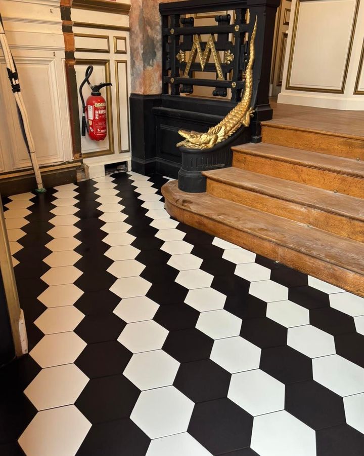 Couloir carrelé en noir et blanc avec escalier en bois, décoration ornée et extincteur rouge fixé au mur