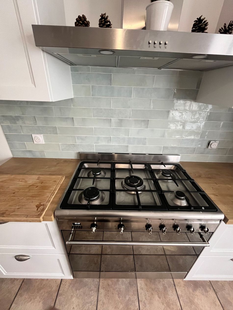 Cuisinière à gaz en acier inoxydable sous une hotte dans une cuisine avec crédence en carrelage bleu pâle et comptoirs en bois.