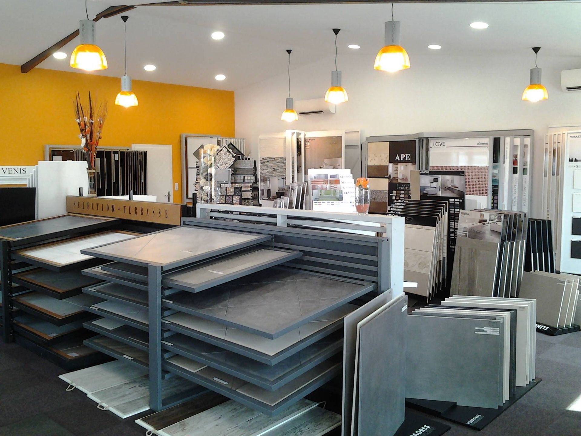 Salle d'exposition de carrelage avec présentoirs de grands carreaux gris sous des suspensions lumineuses.