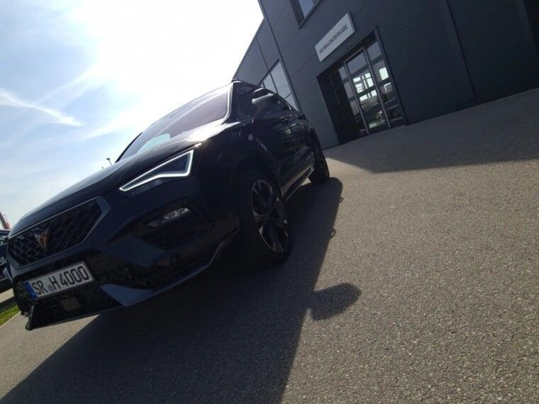 A dark Cupra Ateca SUV parked at an angle in front of a modern commercial building on a sunny day.