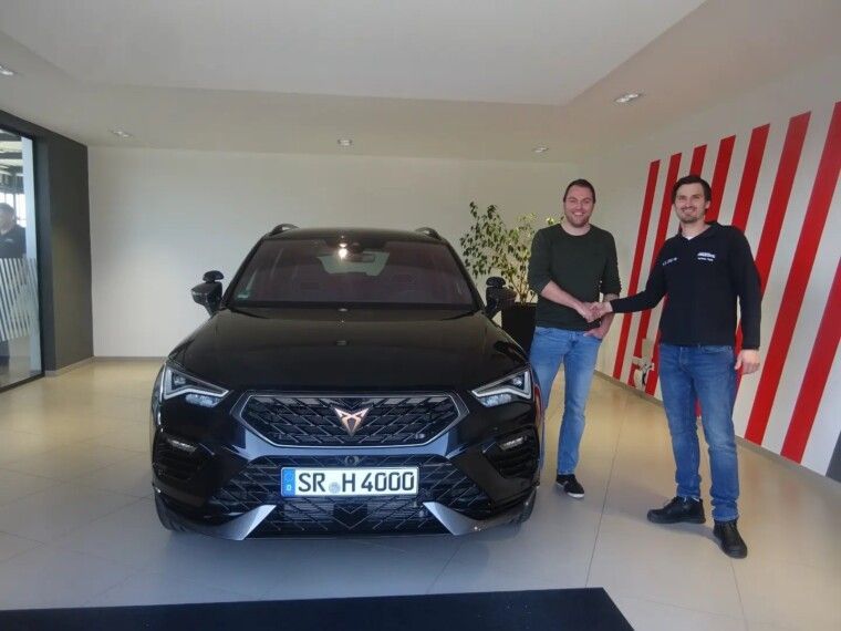 Zwei Personen stehen in einem Autohaus und schütteln sich vor einem schwarzen Cupra SUV mit deutschem Kennzeichen die Hände.