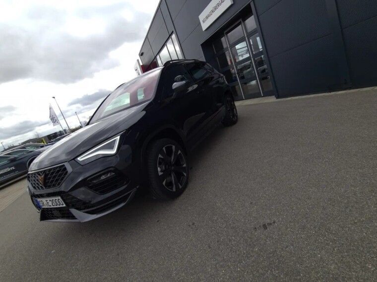 A dark-colored Cupra SUV parked diagonally in front of a modern dealership building under a cloudy sky.