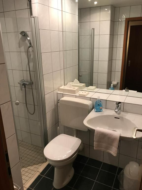 Kleines Badezimmer mit weißen Fliesen. Toilette, Waschbecken, Dusche und Spiegel sind sichtbar.