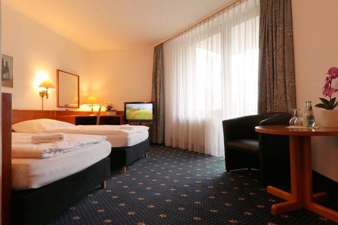 Hotelzimmer mit zwei Betten, Fernseher, Fenster, Sessel und einem kleinen Tisch mit Wasser.