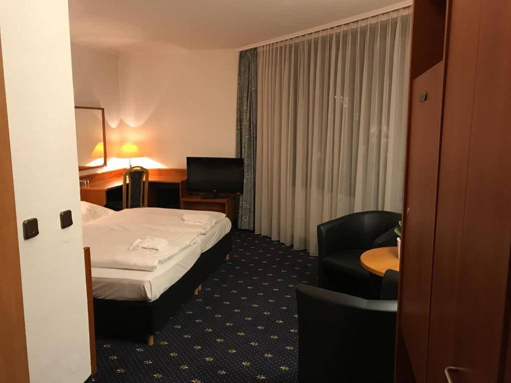 Hotelzimmer mit Bett, Schreibtisch, Fernseher, schwarzen Stühlen und Schrank. Dunkelblauer Teppich, weiße Wände.