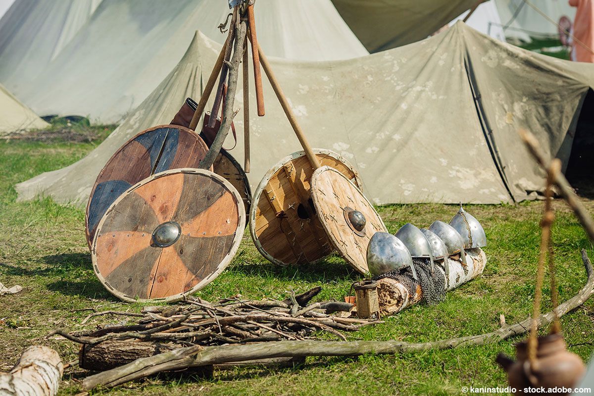 Lagerszene mit Schilden, Helmen und Holzzelten auf grünem Gras.