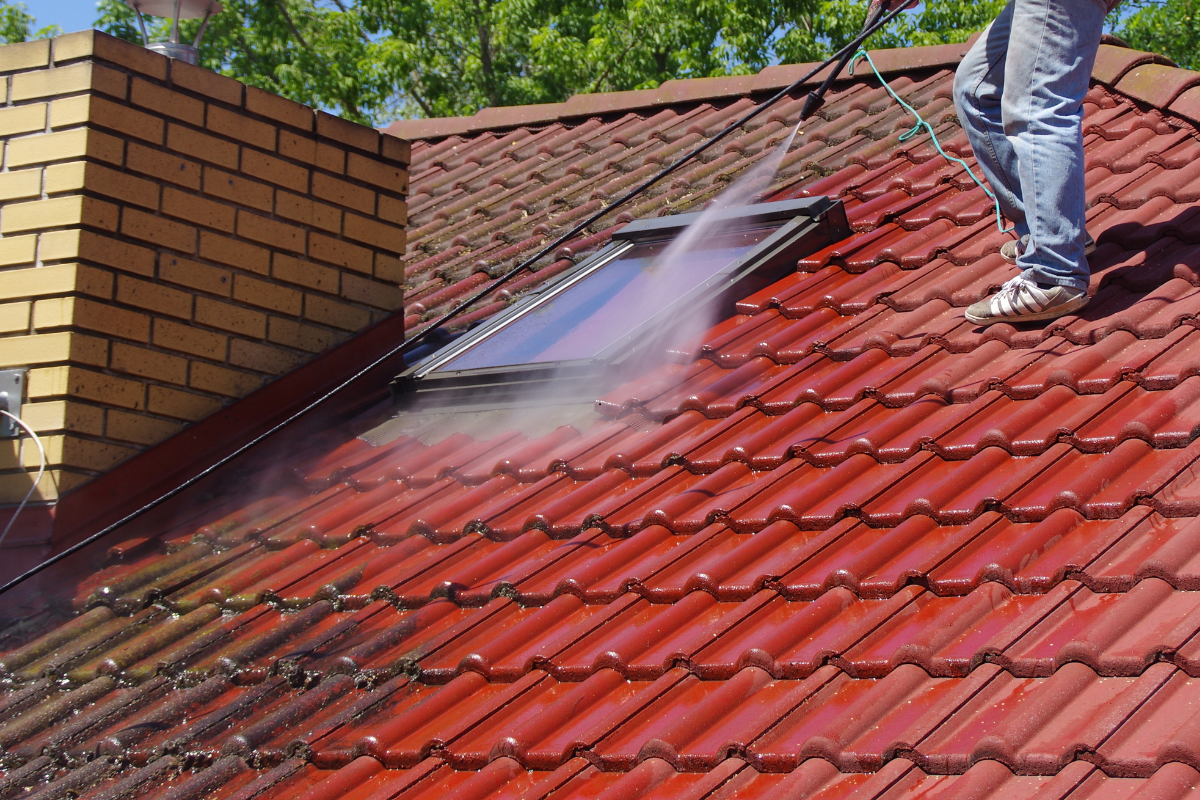 Une personne nettoie à haute pression un toit de tuiles rouges et nettoie la mousse près d'une lucarne.