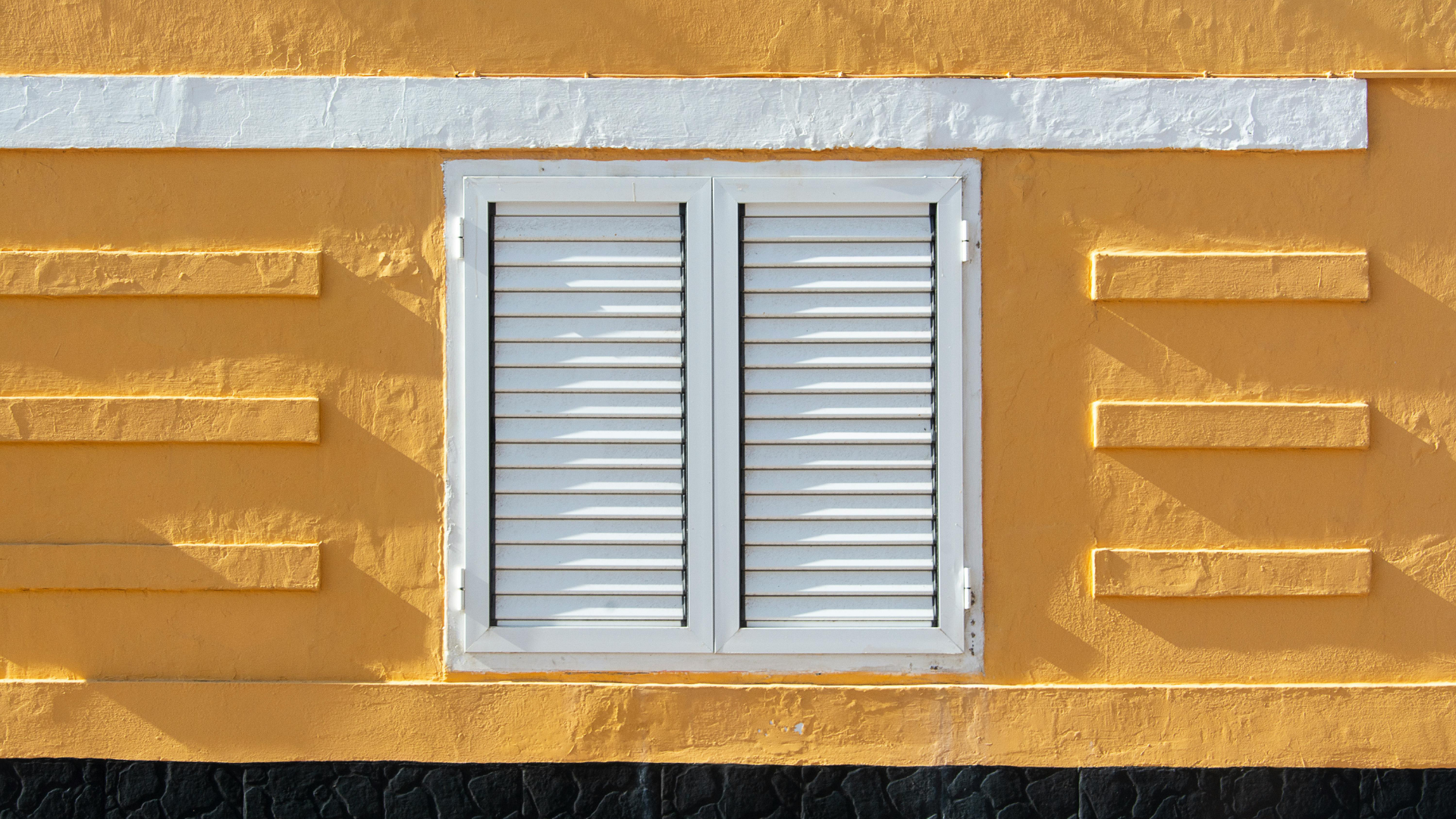 Fenêtre à persiennes blanches insérée dans un mur jaune avec des accents rectangulaires horizontaux et une garniture blanche.