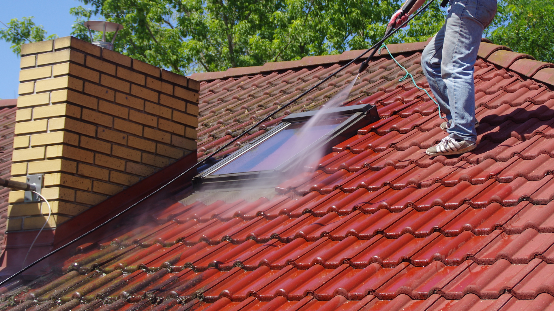 Un homme sur une échelle inspecte un toit de tuiles rouges avec un fleuron décoratif contre un ciel nuageux.
