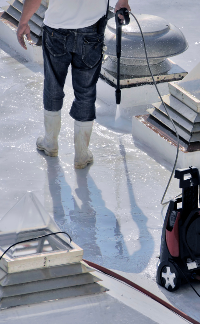 Une personne en bottes nettoie à haute pression un toit commercial blanc à l'aide d'un nettoyeur haute pression.