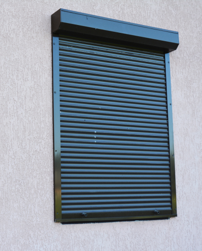 Volet roulant gris foncé recouvrant une fenêtre sur un mur en stuc beige clair.