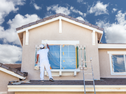 Personne en blanc peignant une fenêtre extérieure sur une maison beige, échelle à côté, fond de ciel bleu.