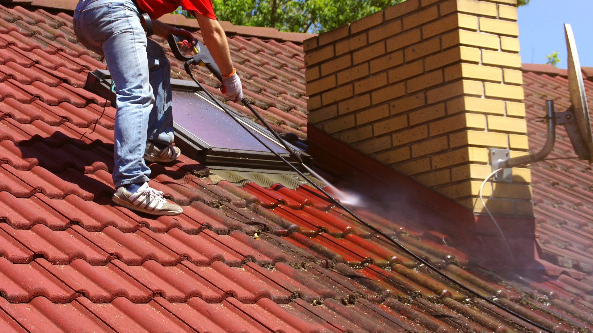 Une personne nettoie à haute pression un toit en tuiles rouges à côté d'une cheminée en briques et d'un puits de lumière.