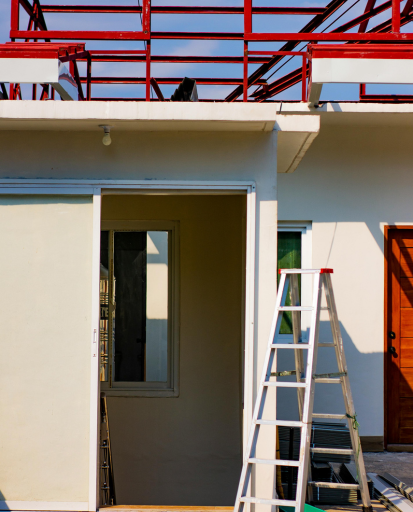 Maison en construction avec charpente métallique rouge, échelle et porte ouverte.