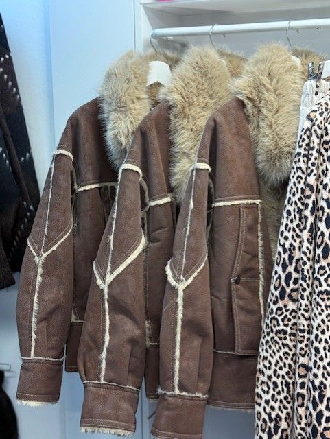 In einem Bekleidungsgeschäft hängen braune Wildlederjacken mit pelzgefütterten Kragen neben einem Mantel mit Leopardenmuster.