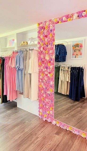 Bekleidungsboutique mit Holzboden und Kleiderständern voller bunter Kleider. Großer Spiegel, umrahmt von rosa Blumen.