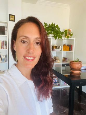 Una persona sonriente con cabello ondulado de color castaño rojizo, vestida con una camisa blanca, se encuentra en una habitación con estanterías y una planta en maceta.