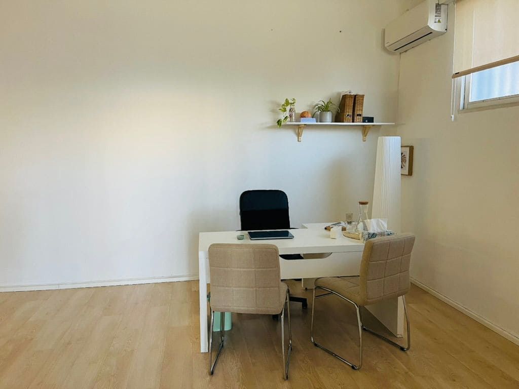 Un escritorio blanco y dos sillas de color beige se encuentran en una habitación luminosa y minimalista con suelo de imitación madera y una pequeña estantería en la pared superior.